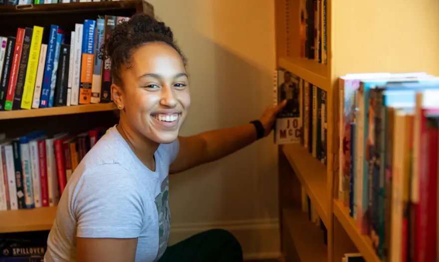 a girl smiles into the camera while reaching for a book on a bookshelf